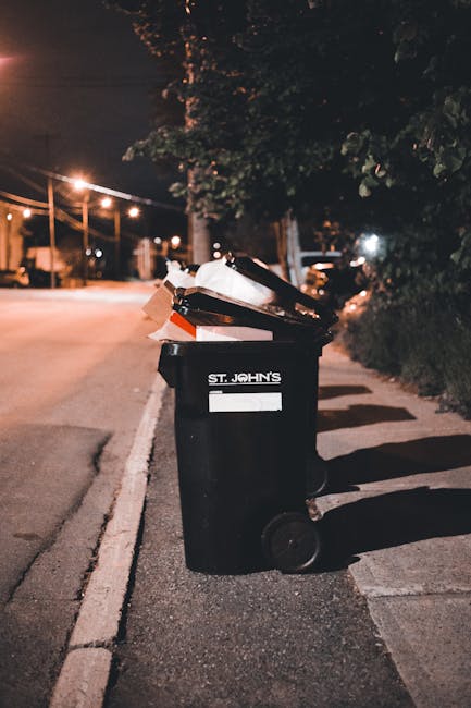 A black wheeled rubbish bin positioned on a sidewalk next to a street at night, with the label 'St. John's' visible on the front. The bin is filled with assorted waste materials, including crumpled pa