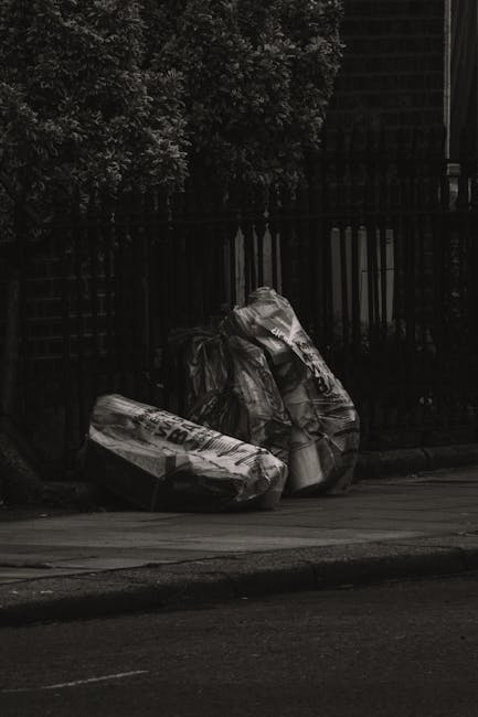 A middle-aged man with dark hair, wearing a black T-shirt with white text and dark trousers, is seen bending forward as he disposes of a white plastic bag filled with rubbish into a tall, stainless st